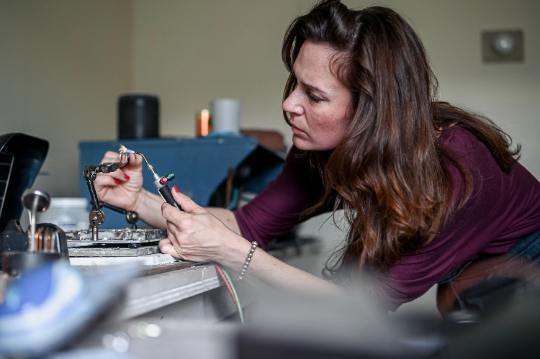 Woman soldering electronic components on a circuit board, focusing intently in a workshop setting adorned with an Emerald Bezel in 14k Gold Necklace.