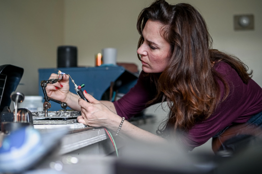 A woman leans over a workbench, soldering electronics with precision as focused as crafting a Custom London Blue Topaz in 14k PD White Gold for Angie.