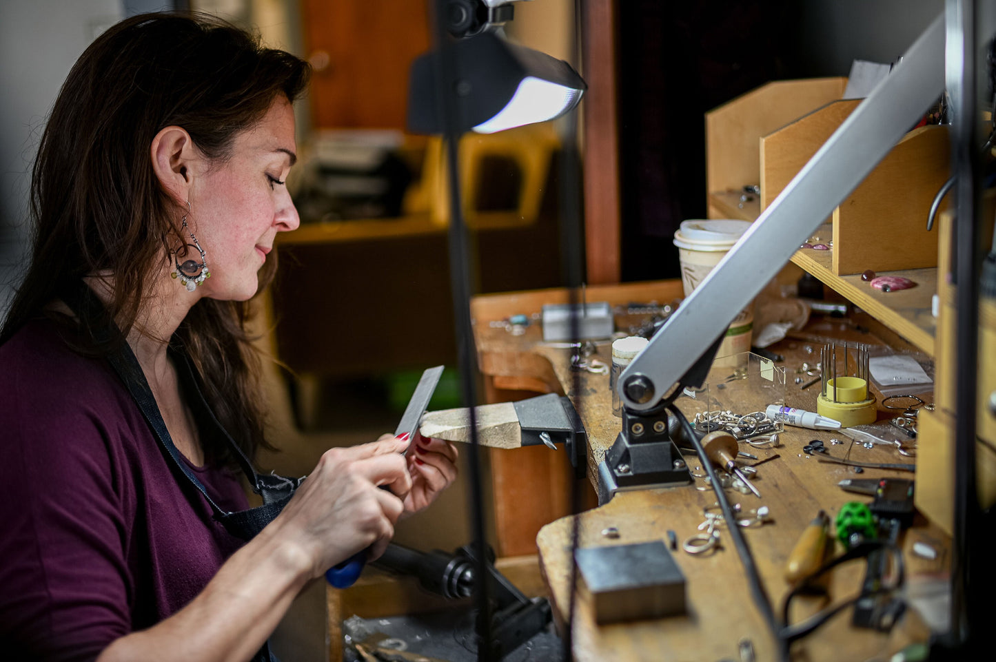 A person sits at a workbench filing metal, surrounded by jewelry-making tools under a desk lamp. Nearby are vibrant turquoise beads ready to be crafted into a unique Custom cut Turquoise and 14k Yellow Gold Ring.