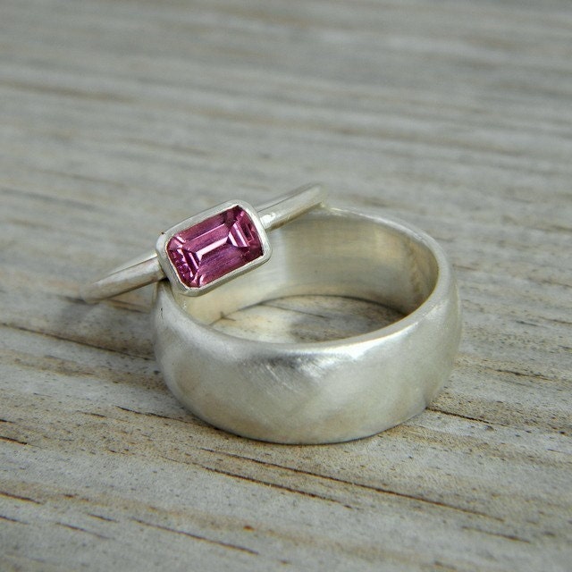 A Pink Emerald Shape Rhodolite Garnet ring, the January birthstone, with a rectangular pink gemstone is set in a thin band and placed inside a thicker plain silver band on a light wooden surface.
