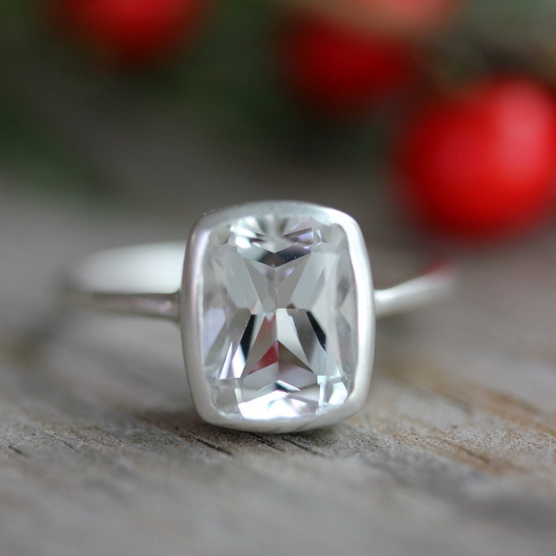 A handmade Cushion White Topaz Ring with red berries in the background, designed by Cassin Jewelry.