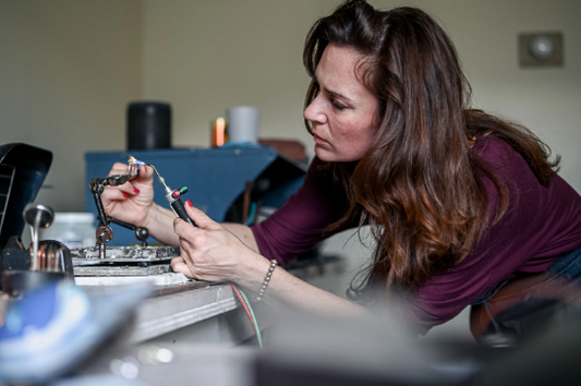 A woman, wearing a Custom Morganite and 14k palladium wedding set, focuses intently on soldering wires at a workstation with various electronic tools.