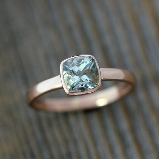 A breathtaking Yellow Gold Aquamarine ring, showcasing a 6mm cushion-shaped light blue gemstone artfully placed on a wooden background, promises ethically sourced elegance.