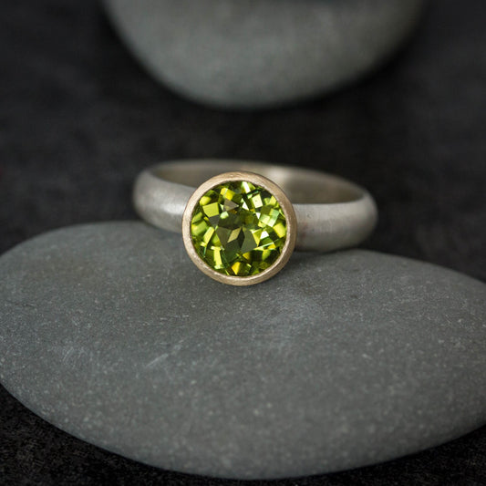 A Handmade Peridot Ring with mixed metals and a peridot stone on top of rocks.