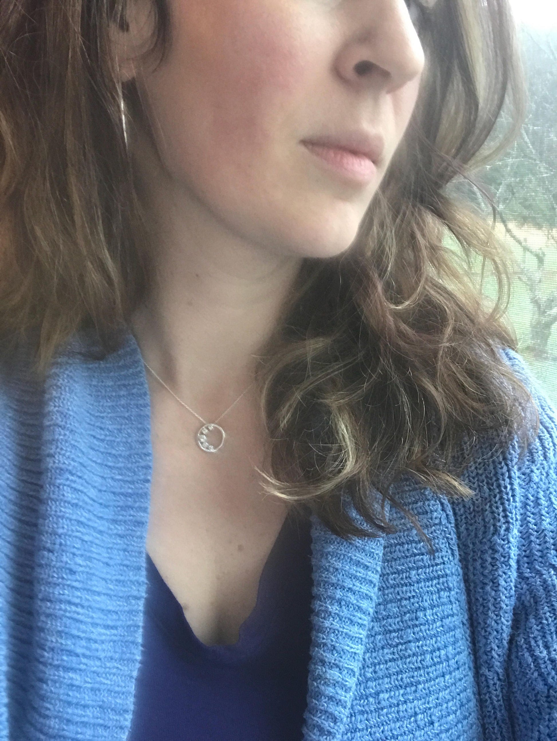 A woman wearing a blue cardigan and a White Sapphire Cresent Moon Pendant Necklace from Cassin Jewelry, known for their handmade jewelry.