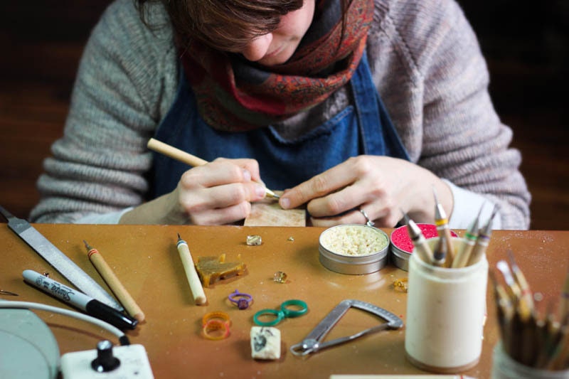 A woman is crafting handmade Moissanite Engagement Rings and Wedding Band Sets at a table with tools.