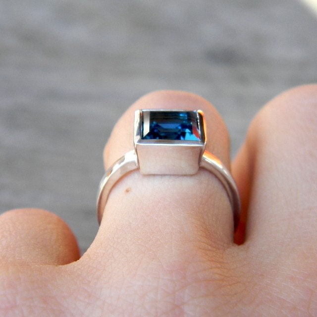 A woman's hand adorned with a stunning cassin jewelry, showcasing a Square London Blue Topaz Ring in handmade Sterling Silver Bezel.