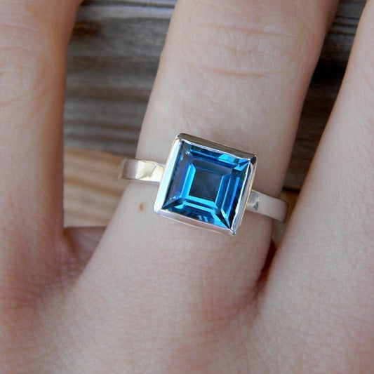 A woman's hand is holding a Square London Blue Topaz Ring, Princess Cut Natural Topaz in Sterling Silver Bezel Bezel by Cassin Jewelry.