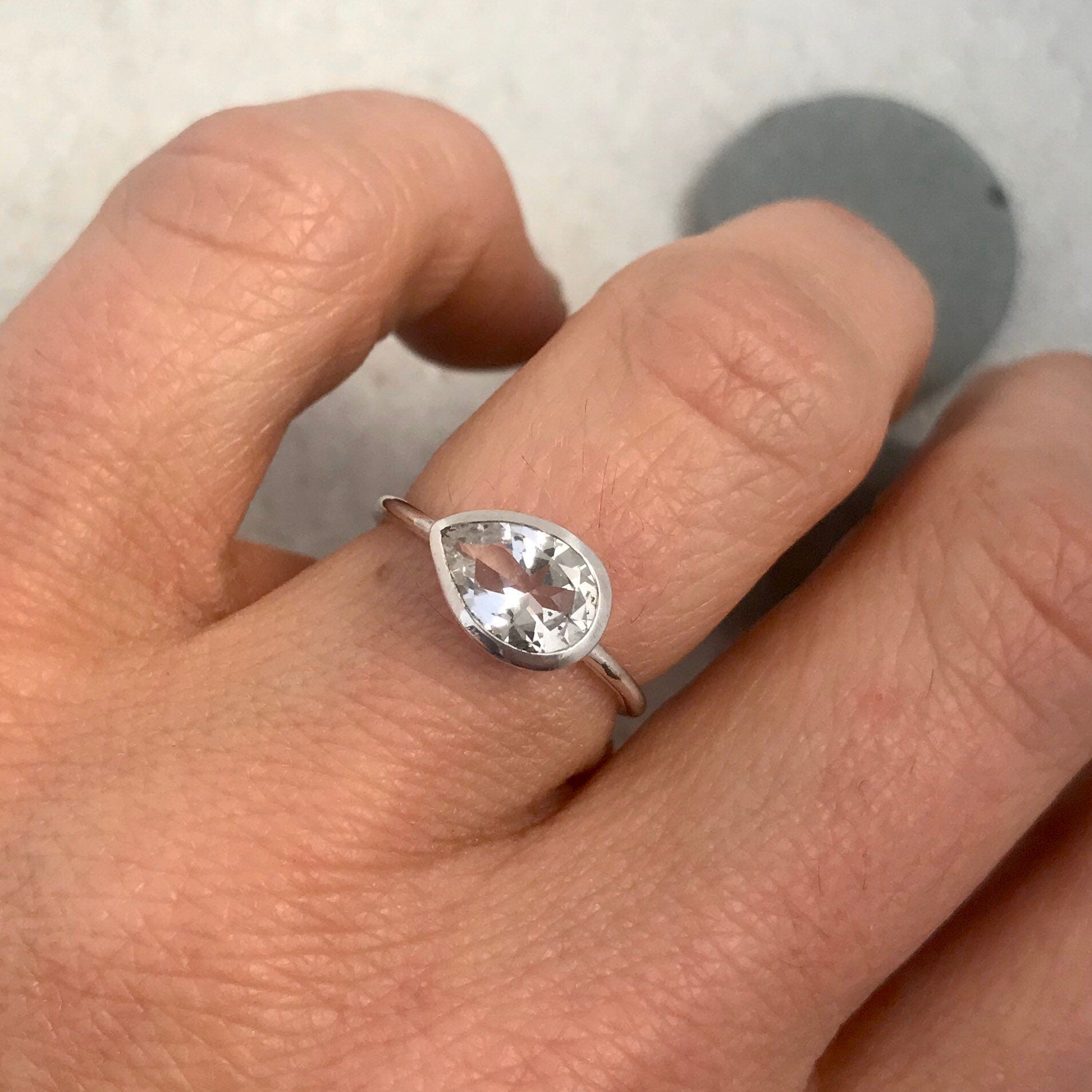 A woman's hand holding a Sideways White Topaz Pear Shape Gemstone Ring, handmade jewelry in Sterling Silver Bezel.