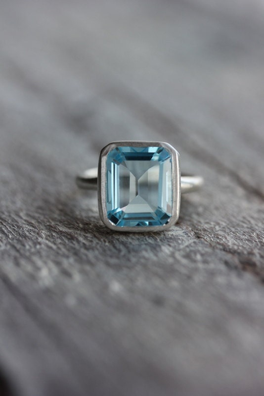 A handmade Sky Blue Topaz Emerald Cut Ring in Recycled Silver on a wooden surface.