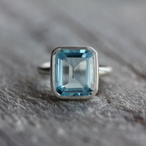 A handmade Sky Blue Topaz Emerald Cut Ring in Recycled Silver on a wooden surface.
