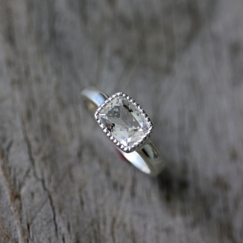 A Handmade Vintage Inspired White Topaz Cushion Cut Ring in Sterling Silver and Miligrain Bezel by Cassin Jewelry on a wooden surface.