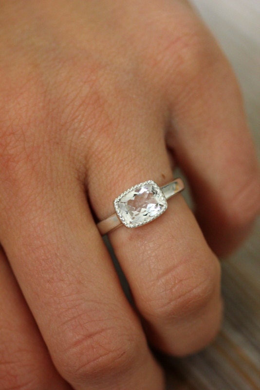 A woman's hand holding a Vintage Inspired White Topaz Cushion Cut Ring in Sterling Silver and Miligrain Bezel engagement ring by Cassin Jewelry.