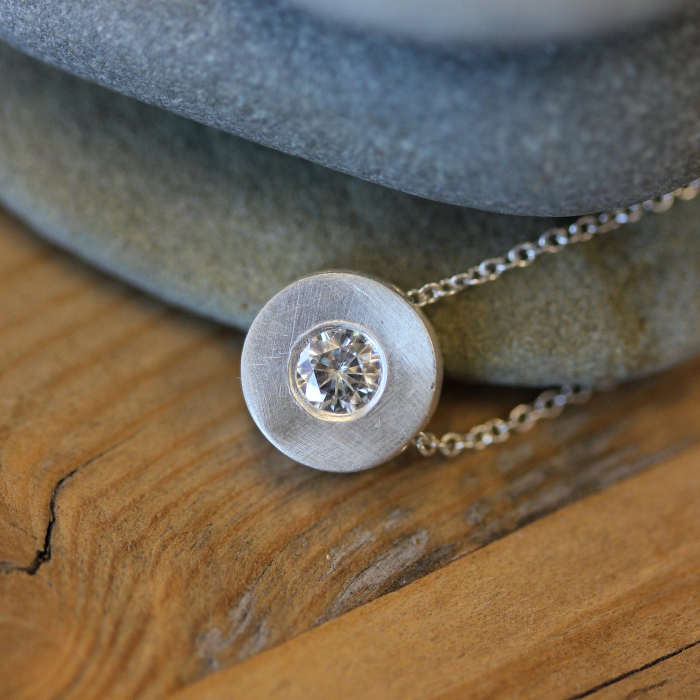 A handmade Moissanite and Sterling Silver Slide Pendant with a diamond on it sits on top of a rock.