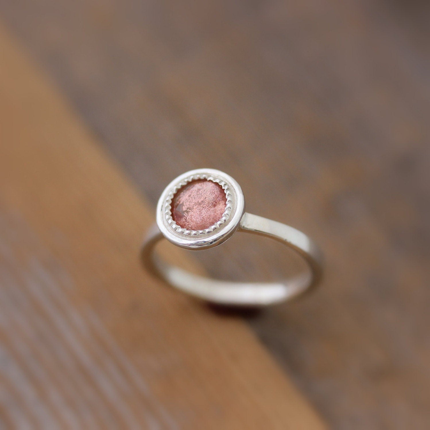 A Handmade Oregon Sunstone Ring in Sterling Silver with a pink stone on top of a wooden table.