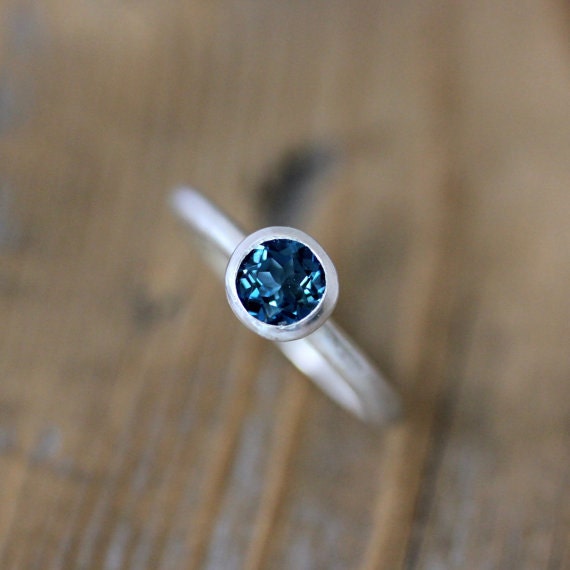 A handmade Cassin Jewelry London Blue Topaz Solitaire Ring on top of a wooden table.