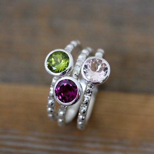 Three handmade Morganite, Peridot and Rhodolite Garnet rings in recycled sterling silver on top of a wooden table.