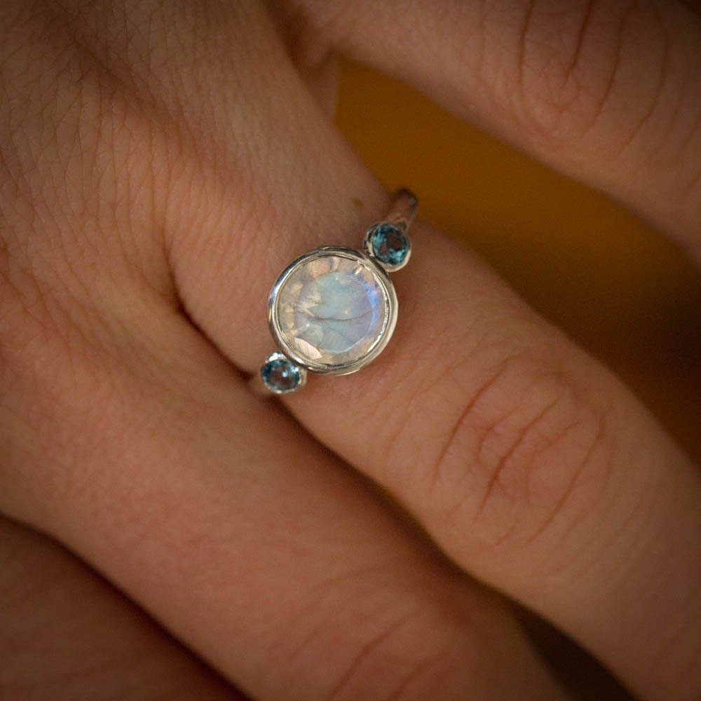 A woman's hand holding a handmade rainbow moonstone and blue topaz ring by Cassin Jewelry.