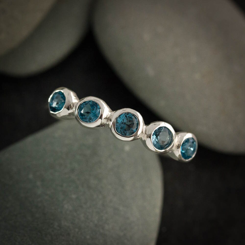 The London Blue Topaz Multistone Ring, featuring five round London blue topaz stones set in a row, is displayed against a background of smooth grey stones.